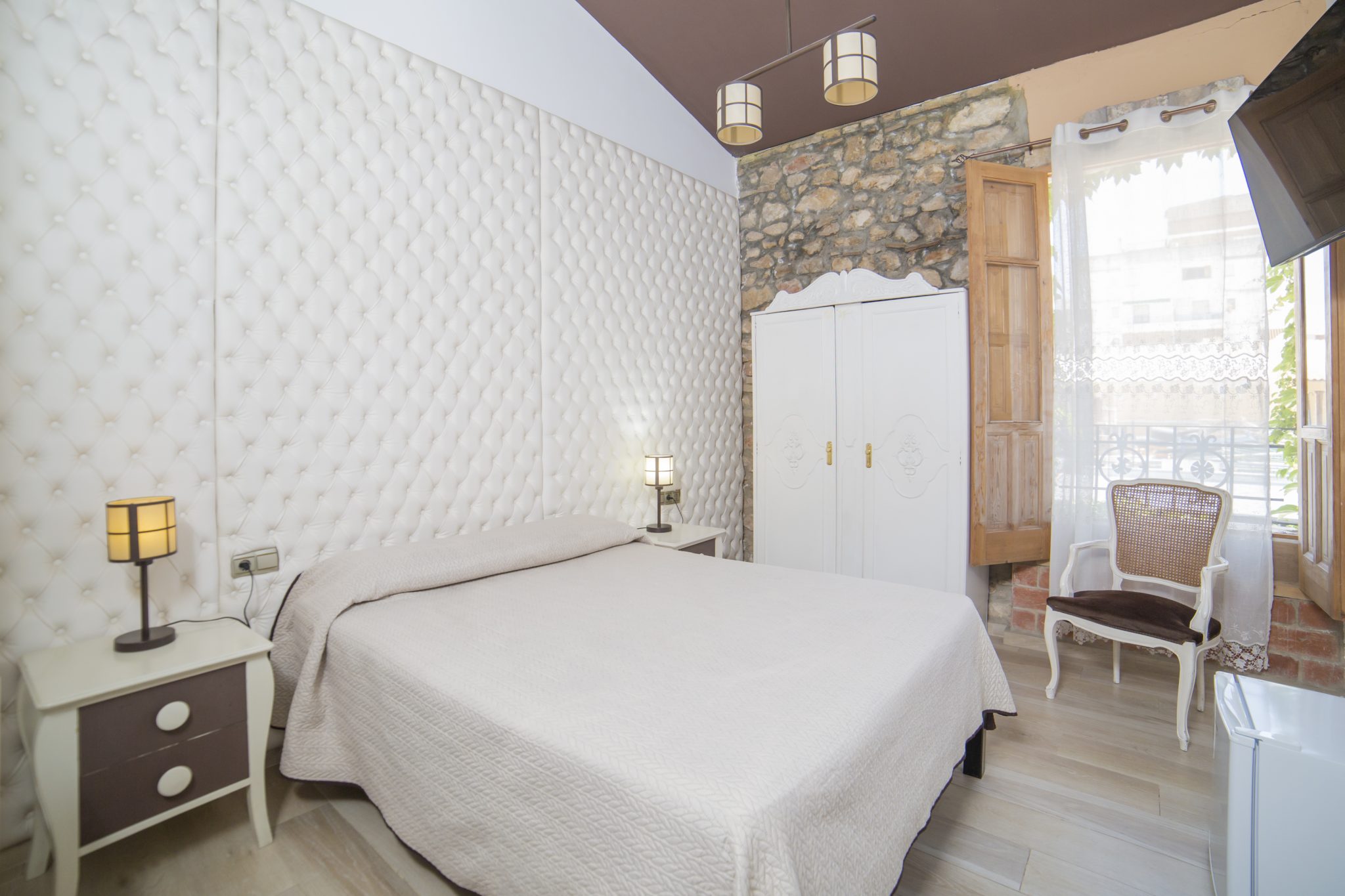 Habitación presidida por una cama de matrimonio, dos mesitas de noche con cajones, pared acolchada, otra pared empedrada donde también se ve un armario grande, una gran ventana abierta que parece dar a un patio, y una silla. También parece apreciarse un minibar.