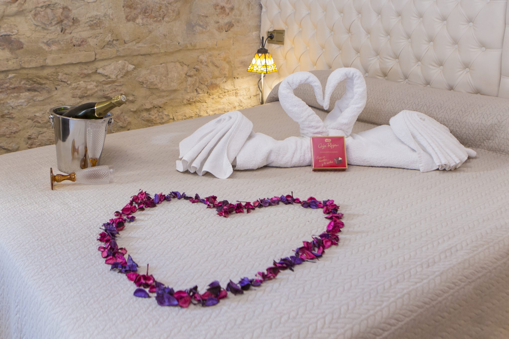 Cama de matrimonio estilizada con un corazón hecho con pétalos de alguna flor, varias toallas en forma de cisnes juntando los cuellos y una botella de alcohol que parece ser cava o champán